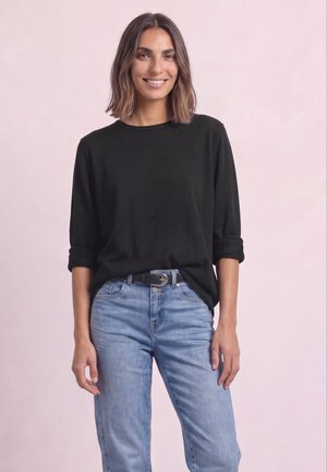 Femme aux cheveux bruns mi-longs souriante, portant une chemise noire à manches longues avec les manches retroussées et un jean bleu clair avec une ceinture noire.