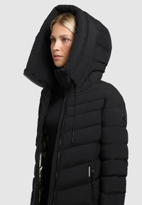 Giacca a vento nera con grande cappuccio, design a cuciture orizzontali e chiusura con zip. Include un distintivo sul braccio sinistro e tasche.