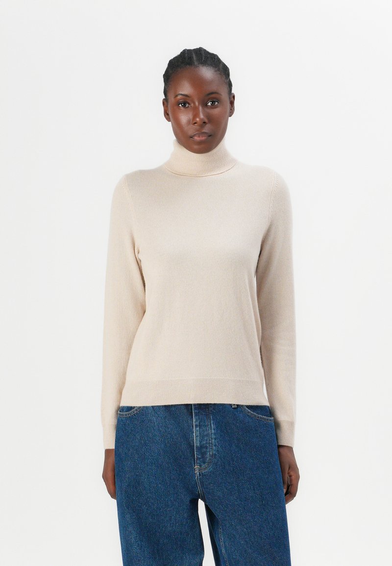 Beige Rollkragensweater aus weichem, strukturiertem Strick, mit langen Ärmeln und schmaler Silhouette, kombiniert mit blauen, hochtaillierten Jeans.