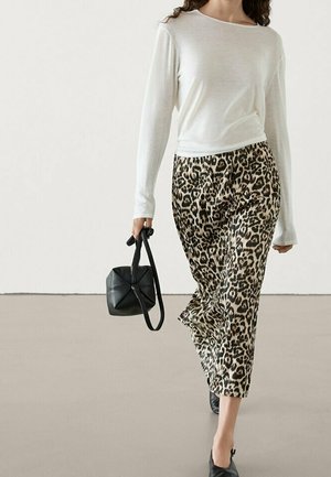 Femme portant un haut blanc à manches longues et un pantalon large imprimé léopard, tenant un sac à main noir, marchant sur un sol uni avec un arrière-plan neutre.