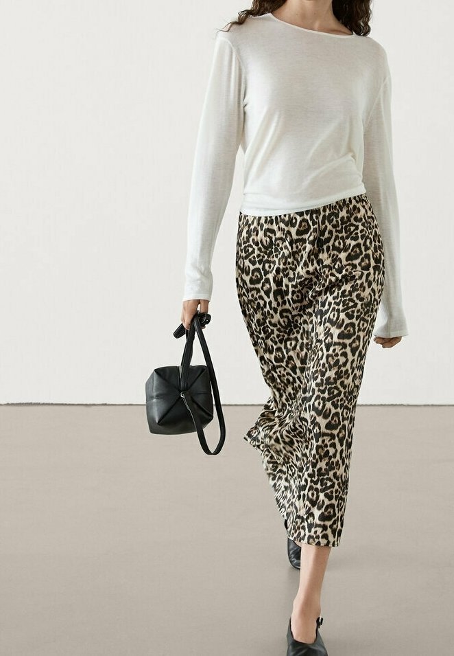 Femme portant un haut blanc à manches longues et un pantalon large imprimé léopard, tenant un sac à main noir, marchant sur un sol uni avec un arrière-plan neutre.