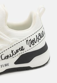 Bijele tenisice s teksturiranim materijalom, crnim potplatom i odvažnim crnim natpisom "Versace Couture" uz stranu, sa šivanim detaljima.