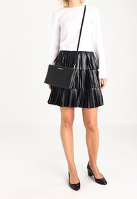 Sort plisseret nederdel med metallisk glans, parret med en hvid langærmet top. En sort crossbody taske med guldbeslag er inkluderet.