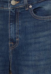 Primo piano della parte anteriore di jeans in denim blu con bottone in metallo e cuciture dettagliate arancioni intorno alla tasca e alla cerniera.