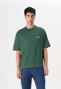 Tricou verde din bumbac, cu mâneci scurte, guler rotund și un mic logo "SELECTED" imprimat pe partea stângă, asortat cu blugi albaștri.