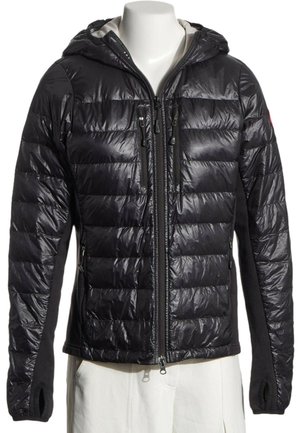 Chaqueta negra acolchada con capucha, cremallera frontal y bolsillos con cremallera en el pecho, mostrada en un torso de maniquí blanco que lleva pantalones blancos.