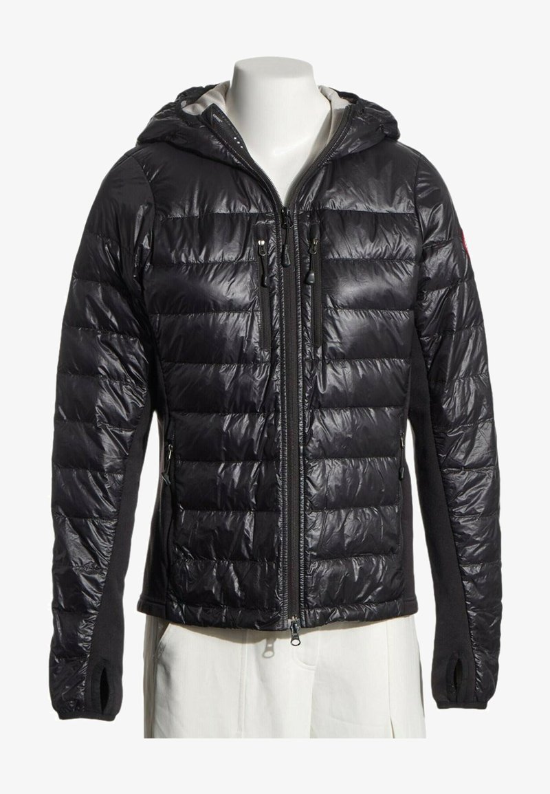 Veste noire matelassée à capuche avec fermeture éclair frontale et poches poitrine zippées, présentée sur un torse de mannequin blanc portant un pantalon blanc.