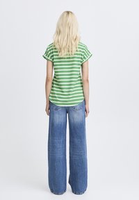 Hosszú szőke hajú nő, zöld-fehér csíkos rövid ujjú felsőt és bő szárú kék farmert visel, hátulról nézve, fehér háttér előtt áll.