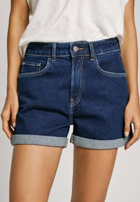 Shorts de mezclilla azul oscuro con dobladillos enrollados, cinco bolsillos y costuras de contraste. Presenta un cierre frontal con botón y un diseño ajustado.