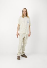 Tricou din bumbac crem cu mânecă scurtă și grafică albastră, asortat cu pantaloni ușor bej și adidași maro cu dungi albe.
