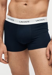 Buste masculin portant un boxer noir avec une ceinture élastique blanche marquée du mot Lacoste en lettres noires.