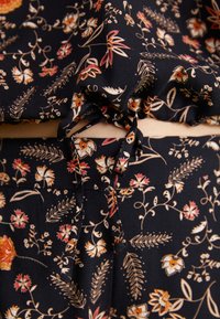 Cordon noué sur un tissu noir avec un motif floral comprenant des fleurs et des feuilles orange, beige et rouges.