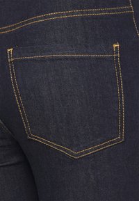 O tecido de denim azul escuro apresenta um bolso traseiro com costura amarela e uma textura subtil. O bolso tem uma forma e design clássicos.