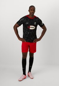 Echipamentul de fotbal al echipei Liverpool FC, care include un tricou negru cu un model și accente roșii, şorturi roșii, șosete negre și ghete roz.