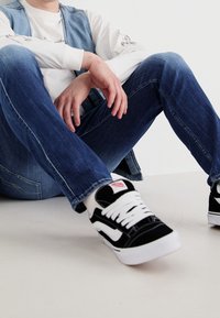 Svarta och vita Vans sneakers med tjocka vita snören, bärs med blå denimjeans och en ljusblå denimväst över en vit skjorta.