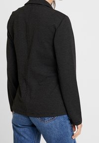 Svart blazer med en subtil vit prickig mönster, strukturerad passform, sjalkrage och långa ärmar, i kombination med blå denimjeans.