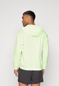 Veste légère à capuche de couleur vert néon éclatant. Comprend des poignets et une taille élastiques, avec une finition lisse et brillante.