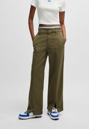 Osoba w wysokich oliwkowozielonych szerokich spodniach z rozcięciami z przodu, białej krótkiej koszulce i niebiesko-białych trampkach, z rękami w kieszeniach.