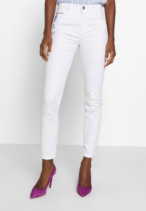 Jean en denim extensible blanc avec une coupe slim, présentant une fermeture avant à bouton et des poches zippées, associés à des talons hauts en daim violets.