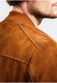 Braune Wildleder-Bomberjacke mit geripptem Kragen, Stickdetails und glatter Textur, die ein modernes Design betont.