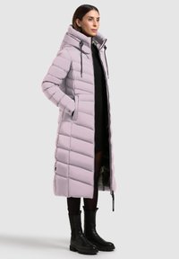 Svetlofialová páperová bunda s dlhým, voľným strihom, prešívaným dizajnom, vysokým golierom, sťahovacími šnúrami a dvoma bočnými vreckami. Prítomné čierne akcenty a čižmy.