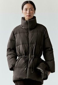 Ciemnobrązowa pikowana kurtka z wysokim kołnierzem, zapinana na zamek z przodu, z sznurkiem w talii. Modelka trzyma czarną, złożoną torebkę. Gładka faktura.