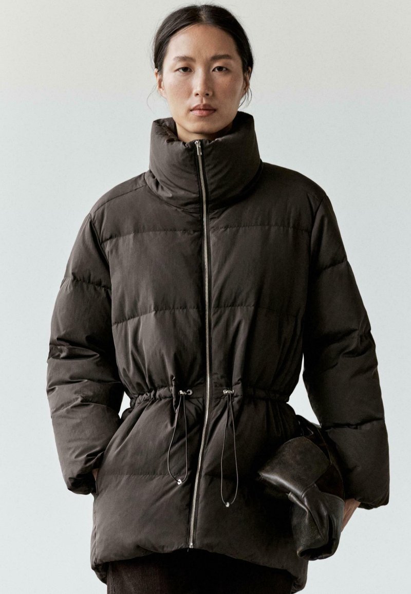 Ciemnobrązowa pikowana kurtka z wysokim kołnierzem, zapinana na zamek z przodu, z sznurkiem w talii. Modelka trzyma czarną, złożoną torebkę. Gładka faktura.