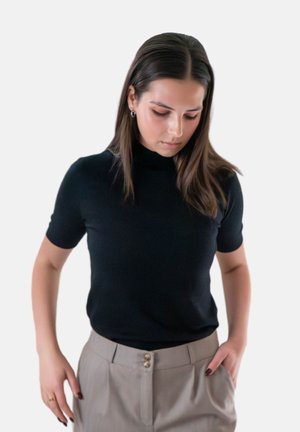 Vrouw met steil bruin haar, gekleed in een zwart t-shirt met korte mouwen en beige hoge taille broek, kijkend naar beneden met handen bij de zakken.