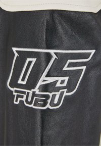 Tissu en cuir noir avec une surface texturée, présentant un logo brodé en blanc, avec des accents crème sur le bord supérieur.