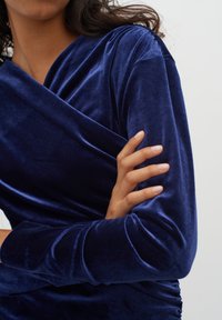 Abito in velluto blu navy con design avvolgente, maniche lunghe e texture arricciata in vita, che mette in risalto la sua morbida lucentezza e struttura fluida.