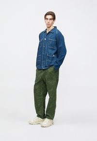 Chaqueta vaquera con dos bolsillos delanteros y costuras blancas, combinada con pantalones anchos de pierna verde texturizada y zapatillas deportivas beige claro.