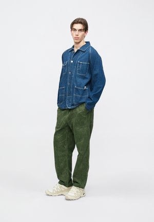 Giacca di denim con due tasche frontali e cuciture bianche, abbinata a pantaloni verdi a gamba larga con trama e sneakers beige chiare.