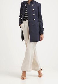 Cappotto navy doppiopetto con bottoni argento, abbinato a una maglietta a righe e pantaloni larghi beige. Sandali completano l'outfit.