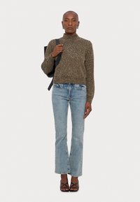 Pull en maille à col roulé dans des tons bruns, jeans taille haute bleu clair, et sandales noires à lanières. Le mannequin tient un sac à dos noir.