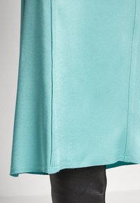 Gros plan sur l'ourlet d'une jupe en satin turquoise porté sur un pantalon en cuir noir, sur un fond neutre.