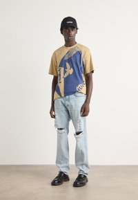 En tan og blå grafisk t-skjorte med fugledesign, parret med lyseblå ripped jeans og svarte flate sko, toppet med en svart caps.