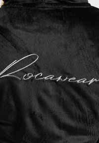 Juodas pūkuotas chalatas su lygiu paviršiumi, ant nugaros siuvinėta sidabrinė "Rocawear" logo. Paprasta dizaino linija su minkštu apdaila.