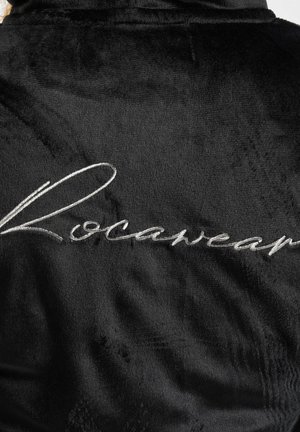 Mustan pörröisen kylpytakin sileämpi pinta, jossa on hopeinen brodeerattu "Rocawear" -logo takana. Yksinkertainen muotoilu pehmeällä viimeistelyllä.