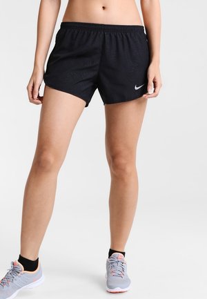 Vrouw die zwarte Nike sportshorts en grijze hardloopschoenen draagt, staat met benen iets uit elkaar op een effen achtergrond.