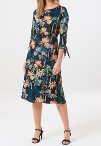 Blumenkleid in Midi-Länge mit schwarzem Hintergrund und bunten tropischen Mustern in Grün, Orange und Blau. Dreiviertelärmel mit Bindebändern.