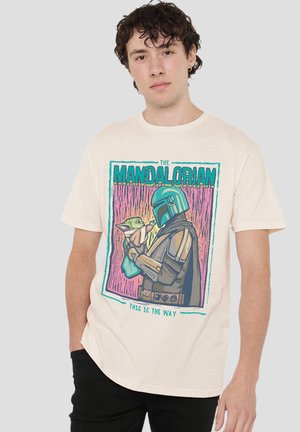Young man wearing cream t-shirt featuring The Mandalorian holding Baby Yoda illustration with text "This is the way."