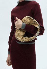 Sac à main marron texturé avec un motif imprimé serpentin et des accents métalliques, tenu par une personne portant une tenue bordeaux avec des poignets gris.