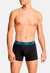 Schwarze Boxershorts mit einem blauen Bund, der das "PUMA"-Logo trägt. Der Stoff wirkt glatt und enganliegend, entworfen für Komfort und Unterstützung.