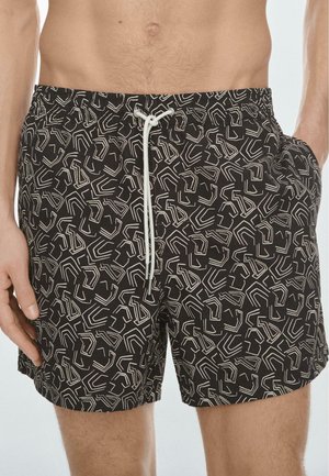 Uomo che indossa pantaloncini da bagno neri con motivi geometrici astratti bianchi e un laccio bianco annodato in vita.