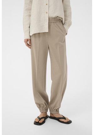 Smėlinės spalvos kelnės su elastine juosmens dalimi ir susmulkintomis kulkšnėmis, dėvimos su šviesia lino marškinėliais su sagomis ir juodomis thong tipo sandalais ant žmogaus.