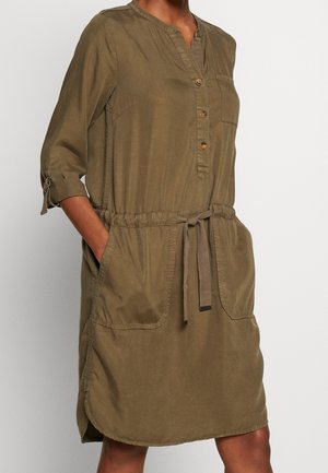 Femme portant une robe boutonnée vert olive, longueur genoux, avec des manches trois-quarts retroussées et une ceinture nouée à la taille, les mains dans les poches avant.