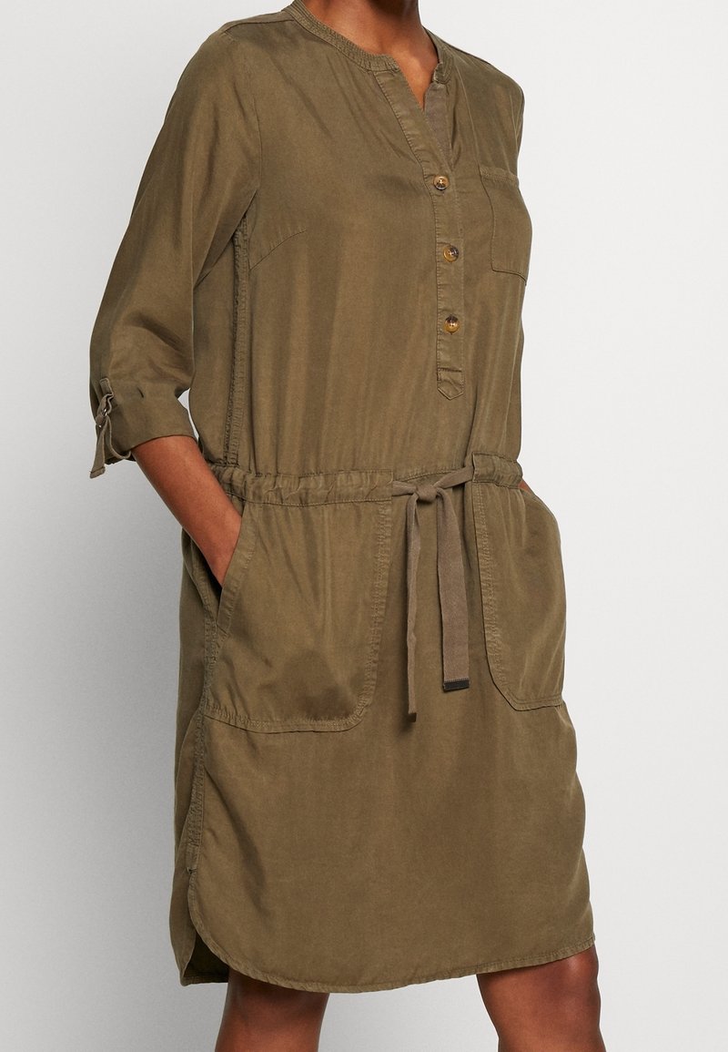 Femme portant une robe boutonnée vert olive, longueur genoux, avec des manches trois-quarts retroussées et une ceinture nouée à la taille, les mains dans les poches avant.