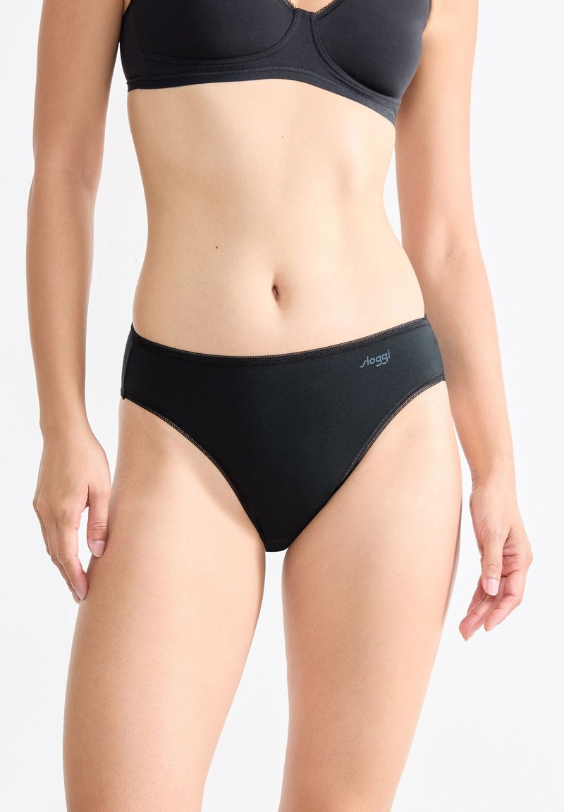 Vrouw in een zwarte katoenen bh en bijpassend zwart bikinistijlondergoed met Sloggi-logo op de heup, staand tegen een effen witte achtergrond.