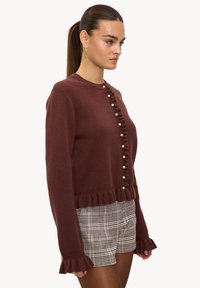 Pull en maille marron avec un col rond, un ourlet et des poignets volantés, et une rangée de boutons nacrés sur le devant. Le mannequin porte un short à carreaux.