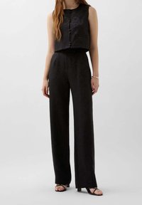 Haut sans manches noir, coupé, avec fermeture à boutons et poche frontale, associé à un pantalon ample taille haute en tissu texturé.